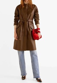 Bruine leren jas met een ceintuur in de taille, zijzakken en een knoopsluiting aan de voorkant. Gecombineerd met lichtblauwe jeans en een rode clutch.