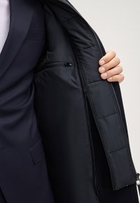 Giacca esterna blu navy con fodera nera isolante. Presenta una texture liscia, tasca interna con zip e design moderno. Taglio aderente.