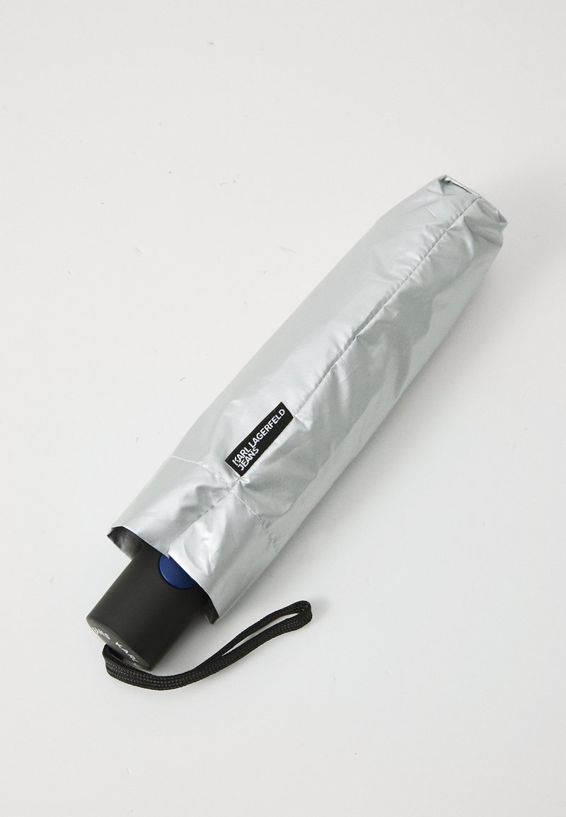 Kompaktiškas sidabrinis skėtis su juodu rankena, mėlynu mygtuku ir audiniu dengiančia viršutine dalimi, ant kurios yra logotipo etiketė. Į komplektą įeina juodas riešo dirželis.