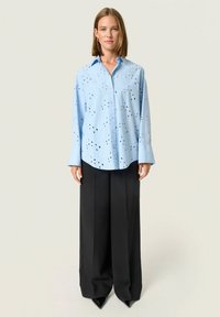 Lichtblauw overhemd met uitsnijdingen, gecombineerd met hoge zwarte wijde broek en puntige zwarte hakken.