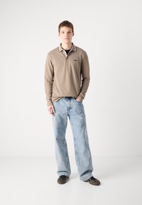 Polo bege de manga longa com um logótipo preto, gola em contraste, combinado com jeans baggy de um azul claro e sapatos escuros, posando sobre um fundo simples.