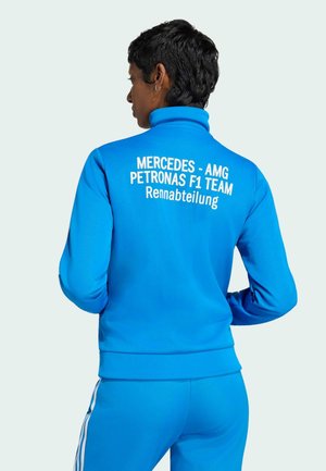 Osoba nosząca jasnoniebieską kurtkę sportową z napisem "MERCEDES - AMG PETRONAS F1 TEAM Rennabteilung" na plecach, stojąca tyłem z rękami na biodrach.