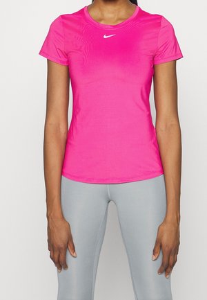 Mujer con camiseta deportiva de manga corta de color rosa brillante con logo blanco y mallas gris claro, de pie sobre un fondo liso.