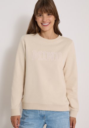 Femme souriante portant un sweat-shirt beige avec le texte "MIND" et un jean bleu clair, debout devant un fond clair uni.