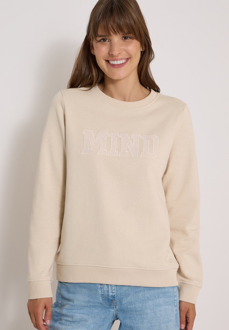 Femme souriante portant un sweat-shirt beige avec le texte "MIND" et un jean bleu clair, debout devant un fond clair uni.