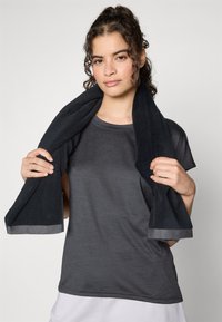 T-shirt sportivo nero in tessuto leggero e liscio con maniche corte. Un asciugamano scuro drappeggiato sulle spalle presenta strisce grigie in accentuazione.