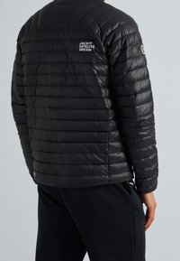 Veste matelassée noire isolante avec coutures horizontales matelassées, col montant et logo sur l'épaule gauche. Texture lisse et brillante.