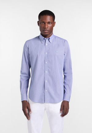 Mann trägt ein hellblaues Hemd mit langen Ärmeln und weißen Hosen, steht vor einem schlichten weißen Hintergrund.