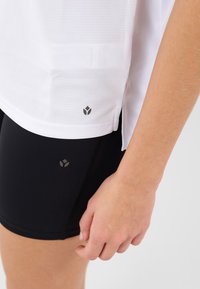 Top sportiv alb, texturat, cu mânecă scurtă și deschidere laterală, asortat cu pantaloni scurți negri, ajustați, cu accente gri ale logo-ului aproape de margine.