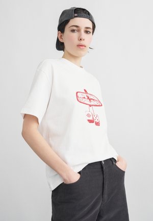 Noor inimene, kes kannab valget T-särki punase küljepeegli ja täringute graafikuga, musti pükse ning tagurpidi halli nokamütsi, käed taskutes.