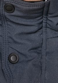 Tela de color gris oscuro con una superficie ligeramente texturizada; presenta dos botones a presión negros y detalles fruncidos a lo largo de la costura.