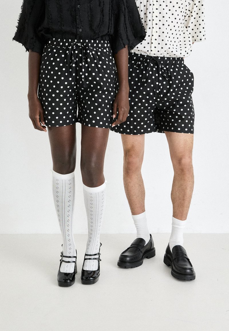 Sorte shorts med hvite prikker på to modeller; den ene har på seg knelengde sokker og svarte sko, den andre har på seg lignende shorts og loafers.