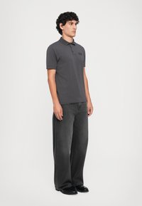 Young man standing, wearing a dark gray polo shirt, loose black jeans, and black shoes, against a plain light background.