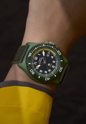 Χέρι με ένα πράσινο ηλιακό ρολόι Timex Expedition με υφασμάτινο λουράκι και ορατό μανίκι κίτρινης ζακέτας.