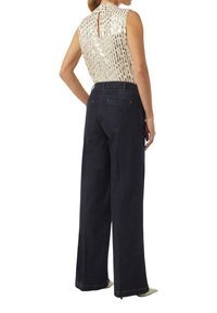 Helles, ärmelloses Oberteil mit Pailletten und einem Schlitz im Rücken, kombiniert mit dunkelblauen, weit geschnittenen Jeans mit Gesäßtaschen und Steppdetails.