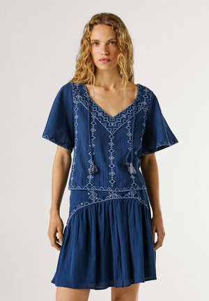 Frau trägt ein blau besticktes Kleid mit kurzen Ärmeln und Quastenbändern, steht vor einem schlichten hellen Hintergrund.