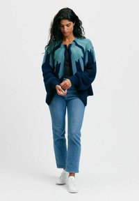 Stickad cardigan med en gradientdesign i teal och mörkblått, med rund halsringning och oversized ärmar, ihop med ljusa jeans och vita skor.