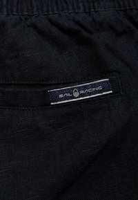 Pantalones cortos de tela negra con cintura elástica, que presentan una pequeña etiqueta con la inscripción "Sail Racing" en blanco y azul. Detalle de superficie texturizada visible.
