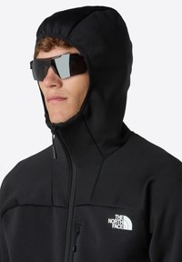 Schwarze, mit Reißverschluss versehene Jacke mit einer taillierten Kapuze, aus glattem Stoff gefertigt. Verfügt über ein Logo auf der Brust und eine Sonnenbrille mit einem eleganten Design.