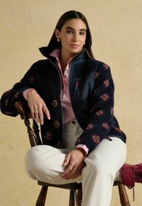 Marineblauwe fleece jas met polka dots, zwarte kraag, ritszak, gedragen over een roze shirt. Model zit op een houten stoel met een witte broek.