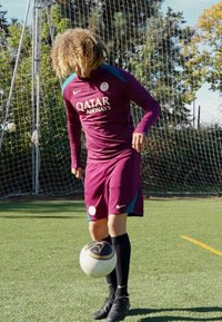 Jalkapalloilija marooninvärisessä Nike-varusteessa sinisin yksityiskohdin, esittämässä temppuja mustavalkoisella pallolla nurmikentällä.