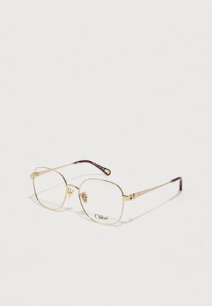 Des lunettes en métal doré avec une monture carrée et des verres transparents, accentuées de couleur marron sur les branches. Nom de la marque "Chloé" visible.