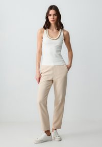 Top bianco a righe con dettagli in catena dorata, abbinato a pantaloni beige e sneakers bianchi. Il modello è in piedi su uno sfondo chiaro.