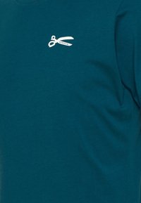 Tyrkysové bavlněné tričko s krátkým rukávem, na levém prsu s malým bílým logem nůžek, hladká textura a kulatý výstřih.