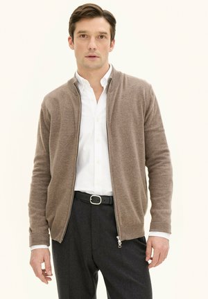 Bruine ritscardigan gemaakt van zacht materiaal, met een geribbelde kraag en manchetten, gecombineerd met een witte knoopshirt en donkere broek.