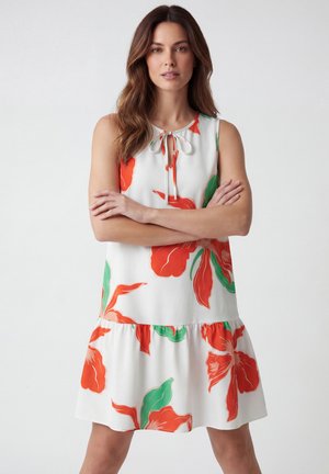 Frau mit langen braunen Haaren, die ein ärmelloses weißes Kleid mit großen roten und grünen Blumenmustern trägt und mit verschränkten Armen steht.