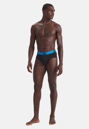 Athletischer Mann, der barfuß steht und schwarze Slips mit einem blauen Bund trägt, auf dem ein schwarzes Adidas-Logo zu sehen ist, und leicht nach links schaut.