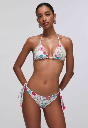 Femme portant un bikini blanc à fleurs rouges, roses et bleues, posant devant un fond gris clair uni.