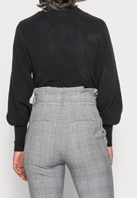 Pull noir côtelé avec manches bouffantes associé à un pantalon gris à carreaux taille haute, doté d'une ceinture et de poches latérales.
