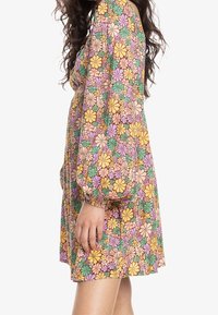 Robe à fleurs présentant un motif multicolore avec des fleurs jaunes, roses et vertes sur un fond beige. La robe a des manches bouffantes et une silhouette cintrée.