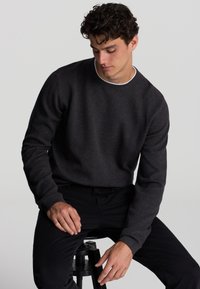 Maglione a maglia grigio scuro con una finitura testurizzata e polsini a coste, dotato di scollatura a girocollo e cuciture minime, abbinato a pantaloni neri.