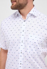 Korte mouw knoopshirt van witte stof, met een blauw patroon van kleine boten en vislijnen, voorzien van een pocket en donkere knopen.