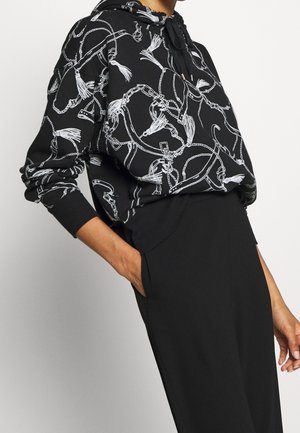 Black hoodie with white tassel and chain pattern worn with black pants, one hand in pocket against white background.