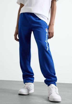 Persoon draagt felblauwe joggingbroek met wit logo in schriftstijl op het been en witte sneakers, staand op een grijze vloer tegen een effen achtergrond.