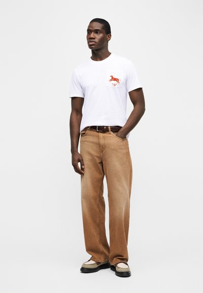 Άνδρας που φοράει λευκό μπλουζάκι με γραφικό άλογο στην τσέπη, καφέ παντελόνι με φαρδιά πόδια, καφέ ζώνη και δίχρωμα παπούτσια, στέκεται μπροστά σε απλό φόντο.