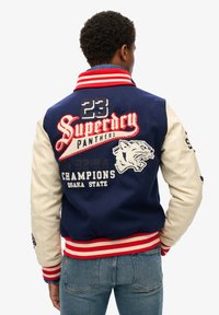 Chaqueta varsity en azul marino y crema con acentos rojos, logotipos bordados y puños y dobladillo de canalé a rayas. Las mangas de cuero en dos tonos mejoran la textura.