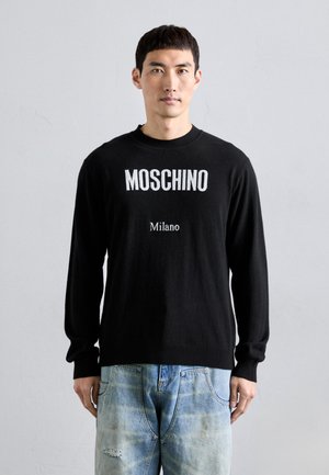 Schwarzer Strickpullover mit dem Schriftzug "MOSCHINO" und "Milano" in Weiß. Langärmlig, mit gerippten Bündchen und Saum, kombiniert mit ausgebleichten blauen Jeans.