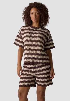 Frau trägt ein zweiteiliges Strickoutfit mit braunen und beigen wellenförmigen Streifen, kurzen Ärmeln und passenden Shorts, steht vor einem schlichten Hintergrund.