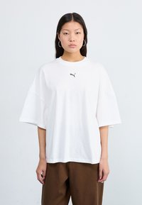 Hvit oversized bomullst-skjorte med korte ermer og rund hals, med en liten svart Puma-logo på brystet. Kombinert med brune bukser.