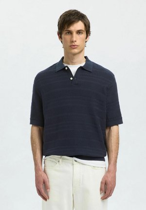 Polo shirt blu navy con un design testurizzato, maniche corte e colletto classico. Presenta tre bottoni e una camicia bianca a strati sotto.