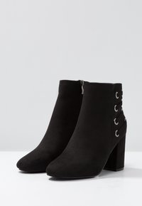 Botas negras hasta el tobillo con una textura de gamuza, punta redonda y una cremallera lateral. Detalle decorativo de cordones con ganchos plateados en el talón.