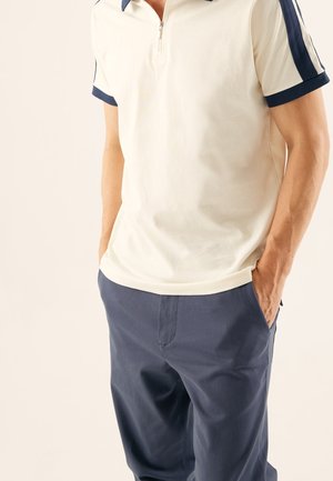 Mann trägt ein weißes kurzärmeliges Poloshirt mit marineblauen Besätzen und Reißverschluss, kombiniert mit marineblauen Hosen, steht mit den Händen in den Taschen.
