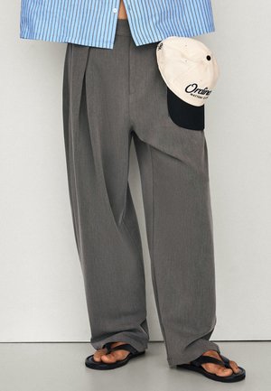 Personne portant un pantalon gris ample, des tongs noires, tenant une casquette beige et noire, avec une partie d'une chemise à rayures bleues visible.