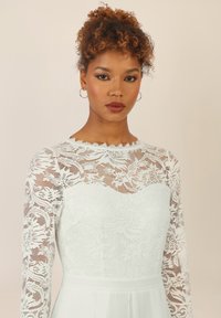 Weißes Spitzenkleid mit langen �Ärmeln, das mit aufwendigen Blumenmustern und einem taillierten Oberteil verziert ist. Glatter Rock, dezenter gewellter Halsauschnitt.