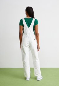 Salopette en coton blanc avec jambes larges et bretelles ajustables, associée à un t-shirt vert. Comprend deux poches arrière et un matériel minimal.