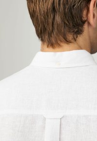 Weißes Leinenhemd mit Kragen- und Knopfdetail auf der Rückseite. Der Stoff hat ein strukturiertes, leichtes Erscheinungsbild, geeignet für warmes Wetter.
